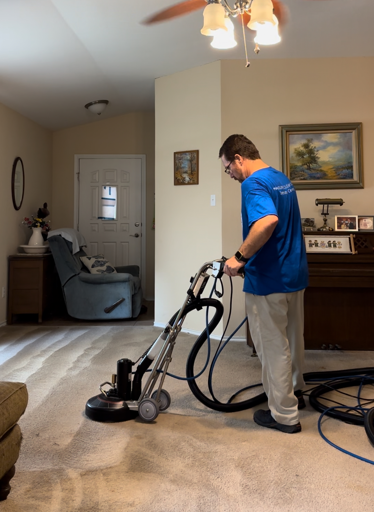 A person wearing yellow gloves is holding the handles of a floor cleaning machine in an office setting, with desks and chairs visible in the background.